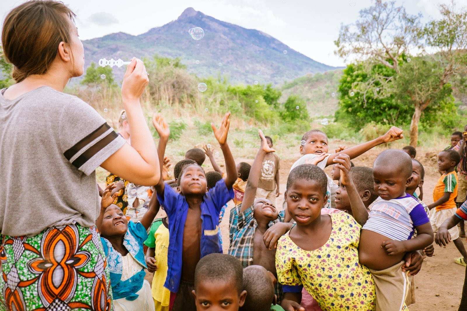 Children reaching for soap bubbles in a village beneath a mountain